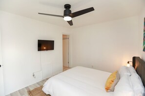 A bedroom with a bed, a ceiling fan, a TV mounted on the wall, and a door leading to another room.