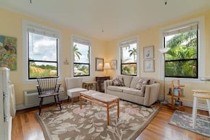A living room with a sofa, armchair, coffee table, and a floral rug. There are large windows with views of greenery.