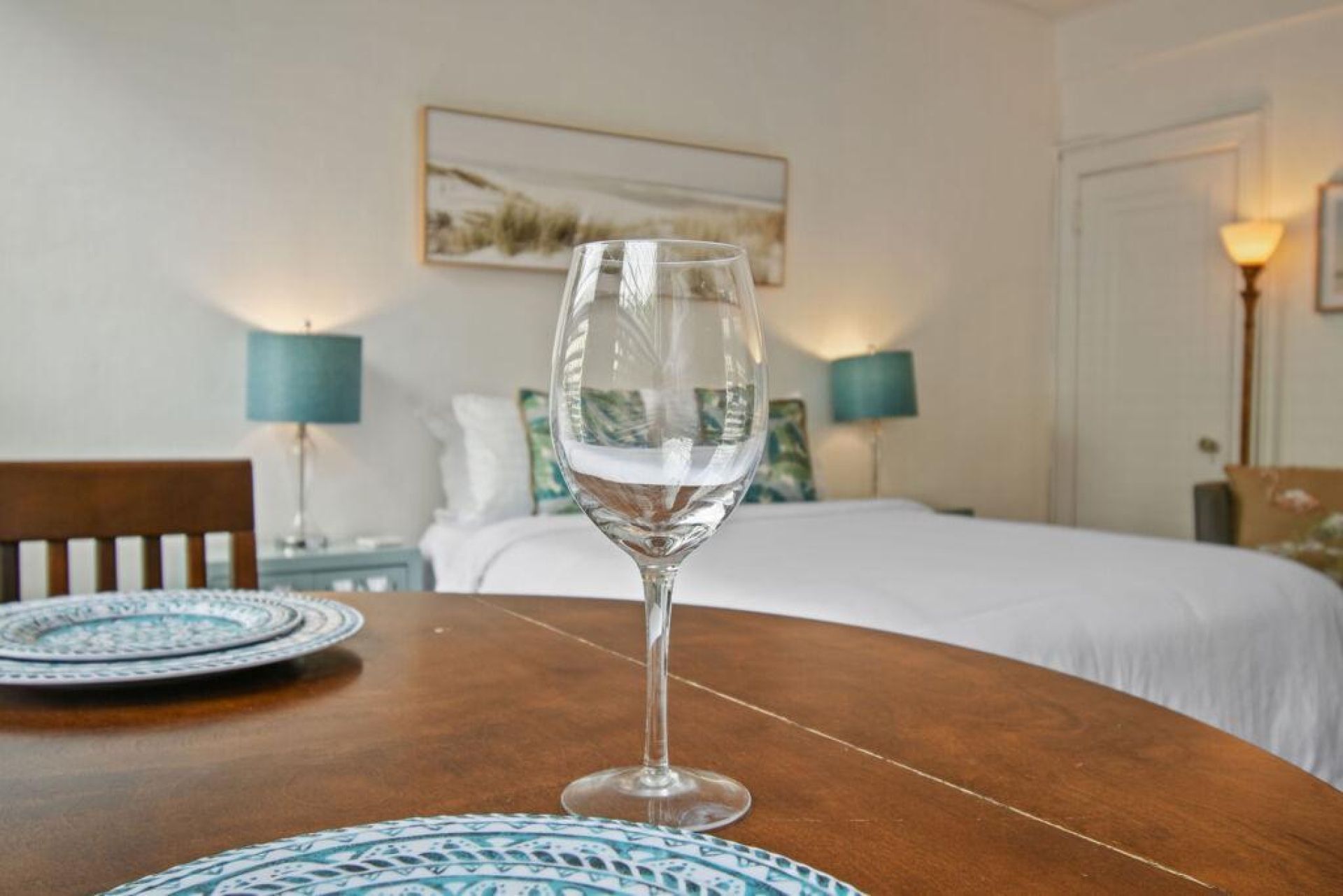 Ein Glas Wasser auf einem Holztisch mit einer Tischmatte mit Muster, ein Bett mit weißer Bettwäsche und ein Bild an der Wand.