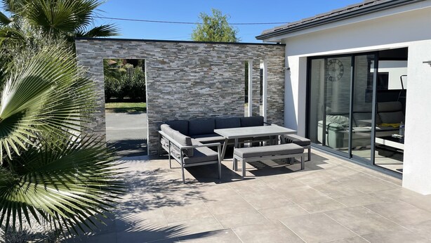 A modern outdoor patio with a stone wall, a grey sofa, and a table. - Magnificent contemporary property with heated spa 15 min from Bordeaux center. (Fargues-Saint-Hilaire)