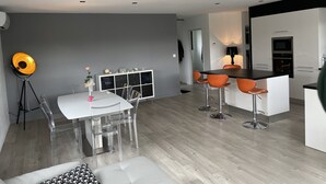 Una cocina moderna con armarios blancos, encimera negra y horno integrado. Hay una zona de comedor con mesa blanca y sillas transparentes.