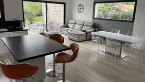 Una cocina moderna con comedor, sofá y un amplio ventanal con vistas a vegetación y una casa.