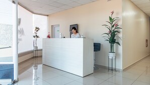 Een receptie met een wit aanmeldadres, een receptionist, een potplant en een deur met een bordje.