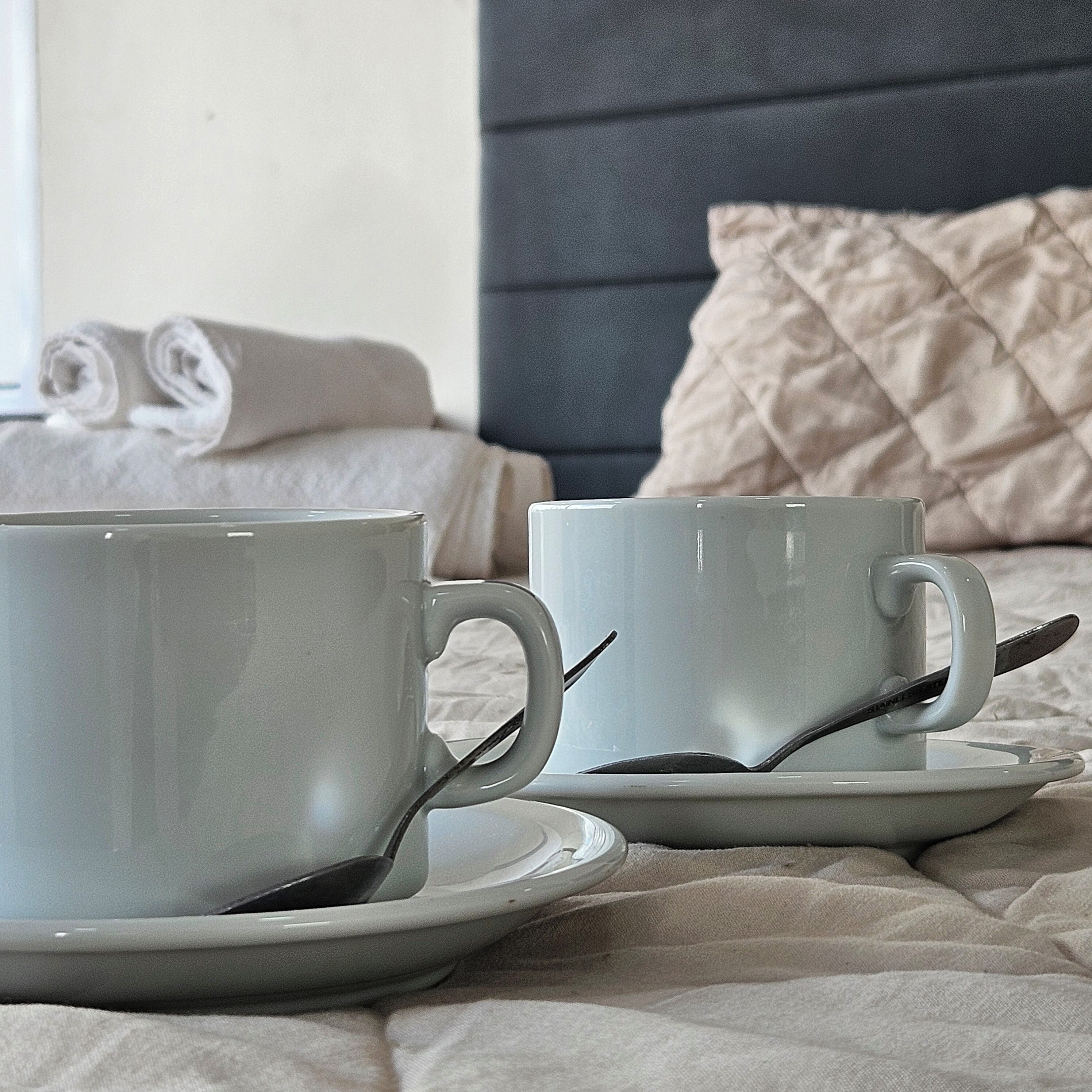 Two white cups on a bed with a grey headboard and a beige quilted pillow.