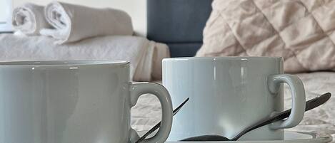 Two white cups on a bed with a grey headboard and a beige quilted pillow.