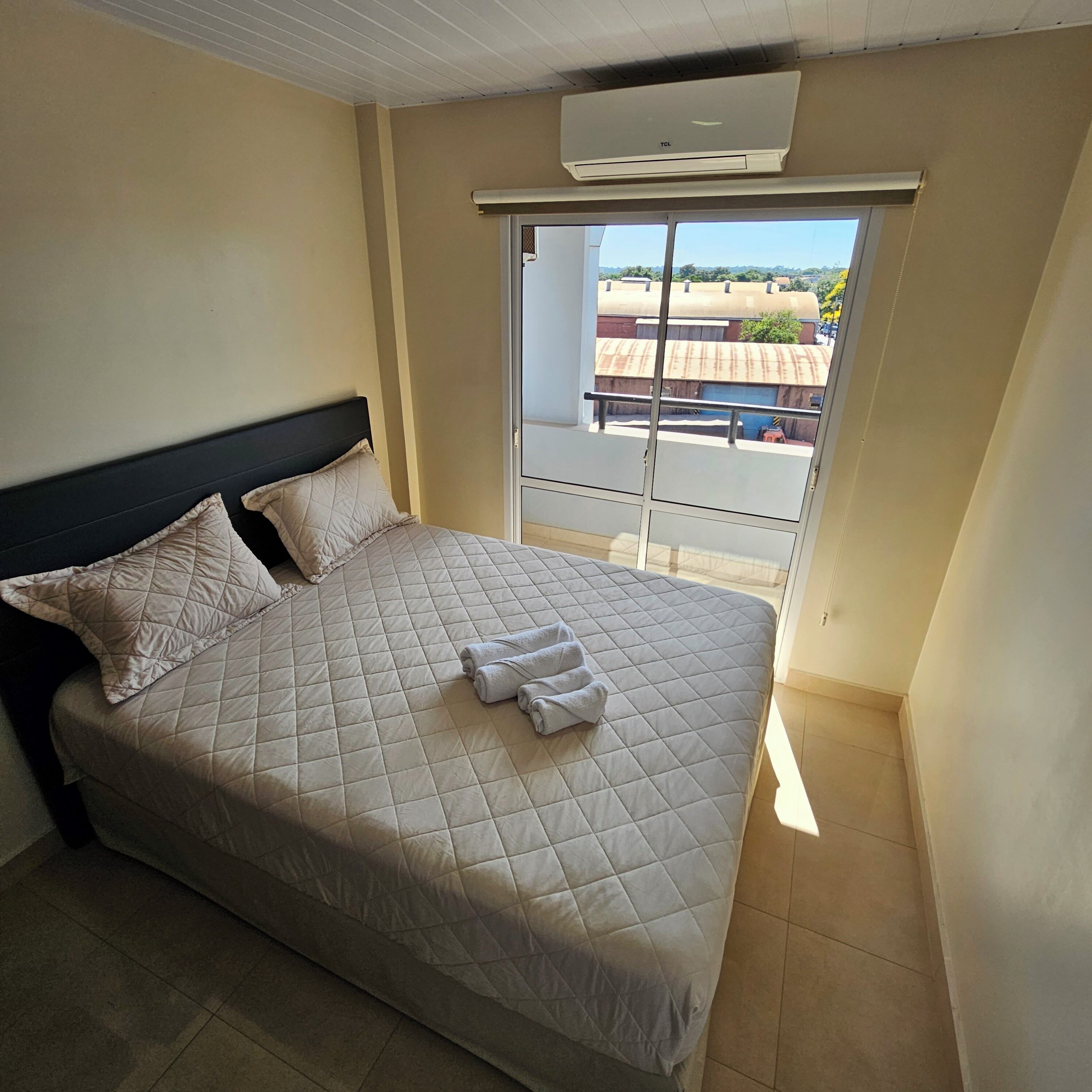 A bedroom with a bed, pillows, a rolled towel, an air conditioning unit, and a view of buildings through a window.