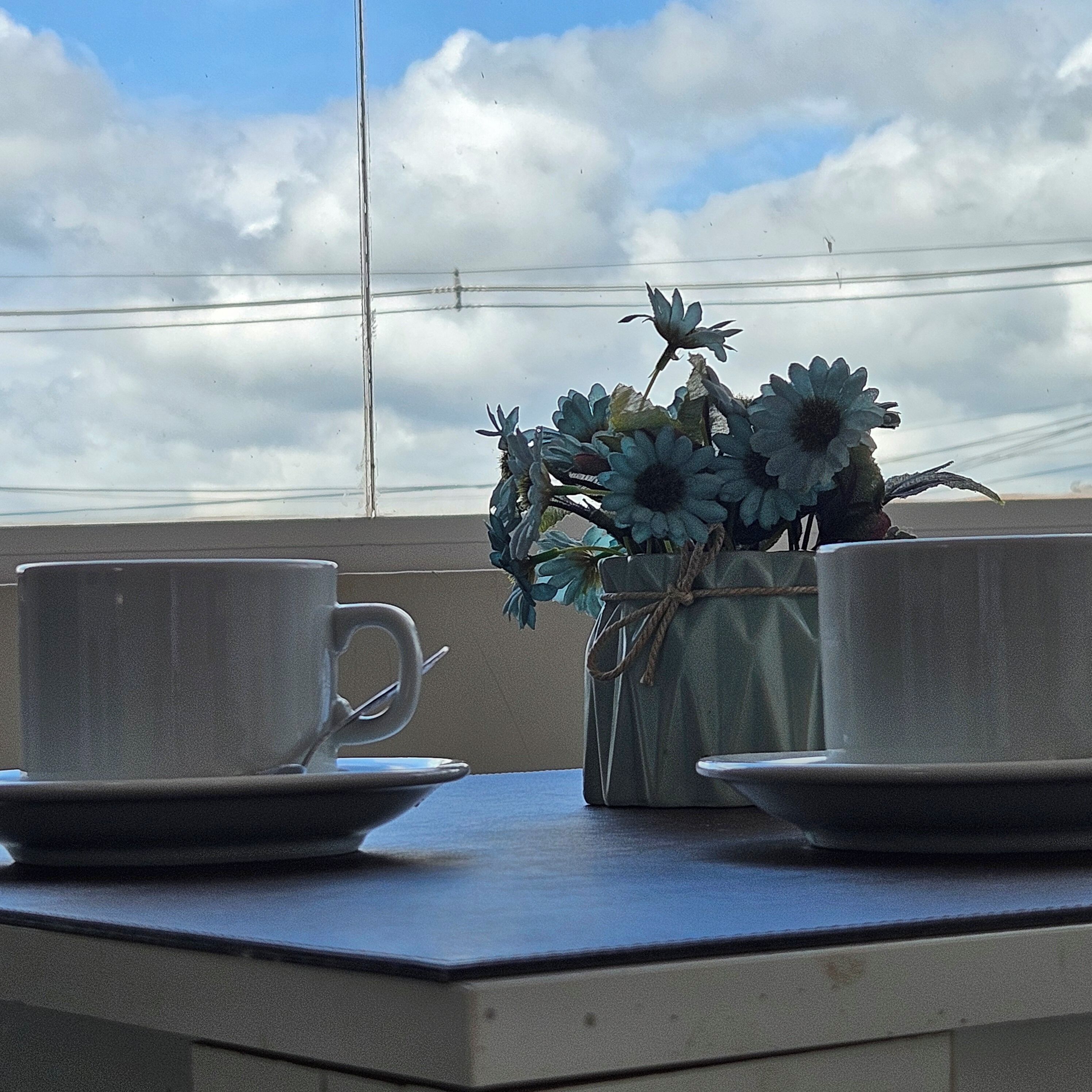 Taula amb dues tasses i platets, un ram de flors artificials i vista del cel amb núvols.