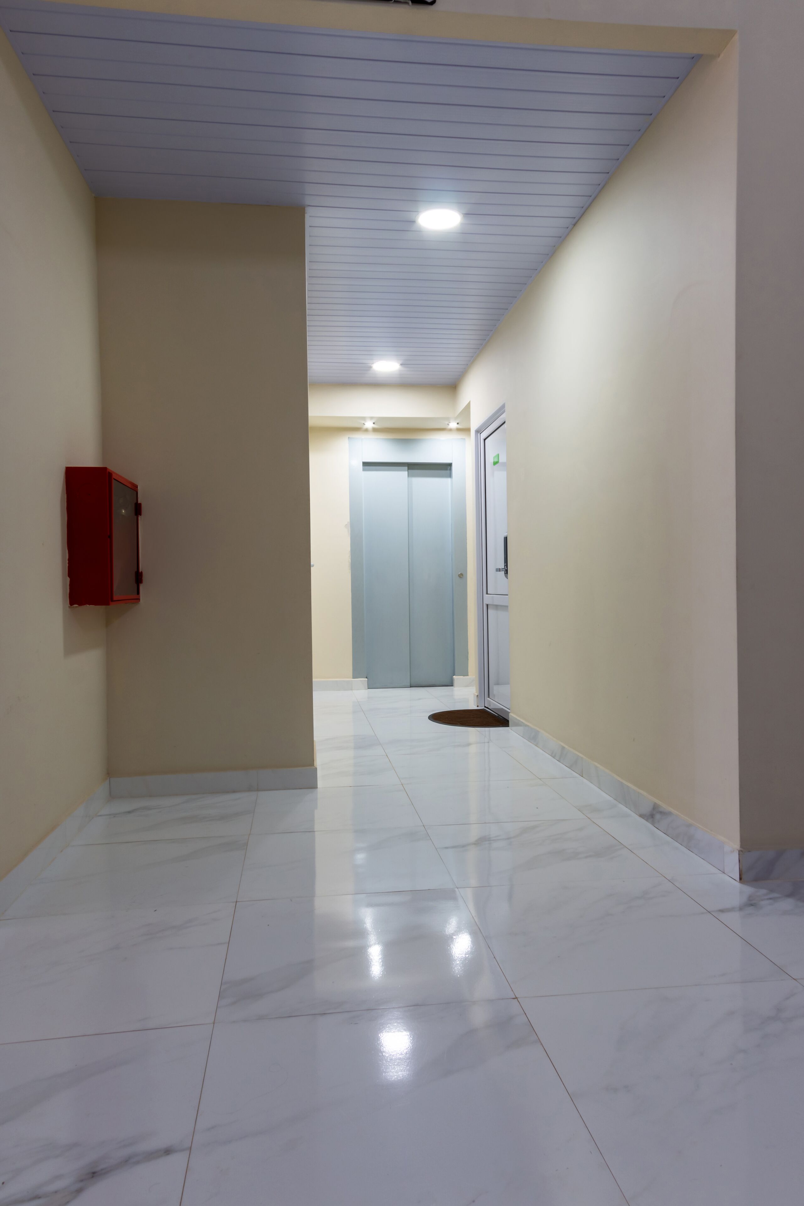 A hallway with a red fire extinguisher, a door, and a light fixture on the ceiling.