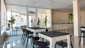 Een moderne, goed verlichte cafetaria met witte tafels en zwarte stoelen, potplanten en een duidelijk zichtbaar vluchtroutebord.