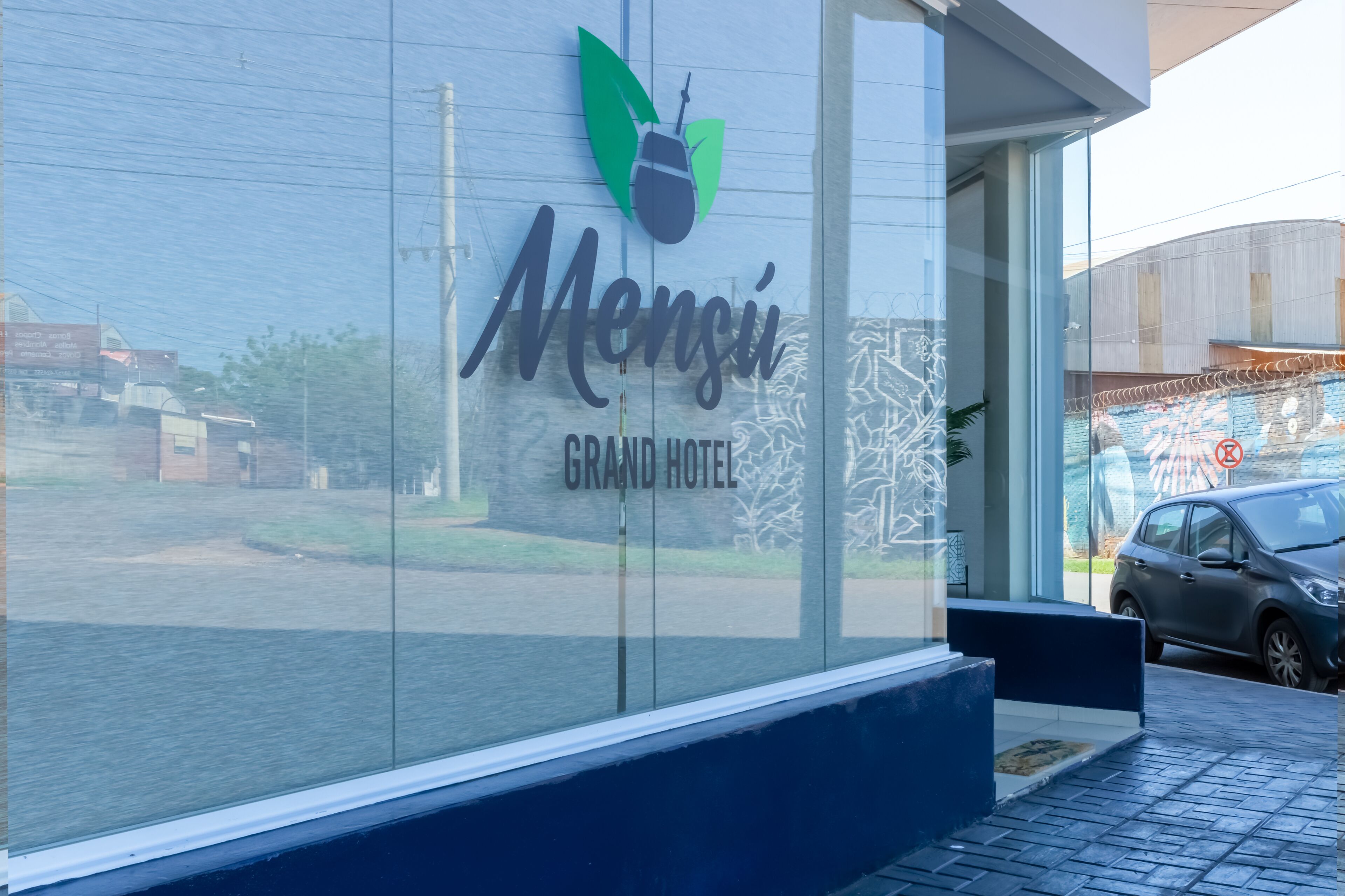 The exterior of the Mensi Grand Hotel with a clear glass facade, a logo featuring a beetle and leaves, and a view of the street and parked cars.