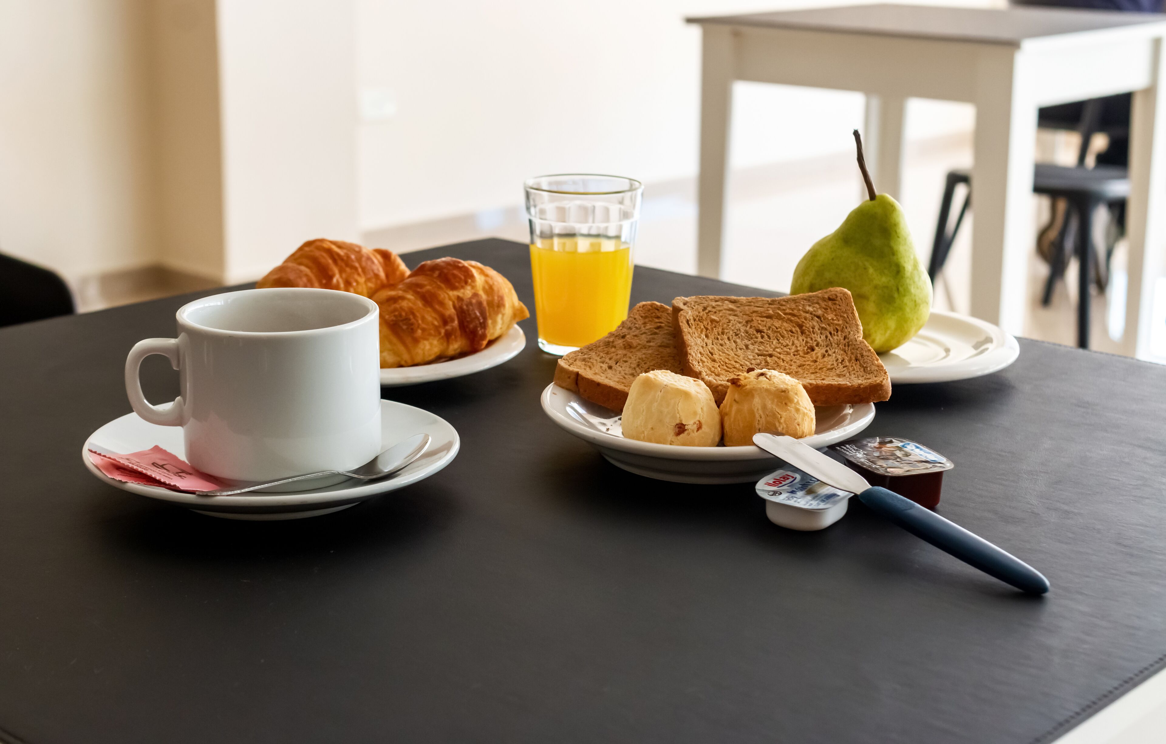 Desayuno con croissants, tostadas, una pera, jugo de naranja y una taza de café sobre una mesa.