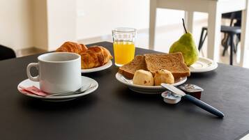 Desayuno con croissants, tostadas, una pera, jugo de naranja y una taza de café sobre una mesa.