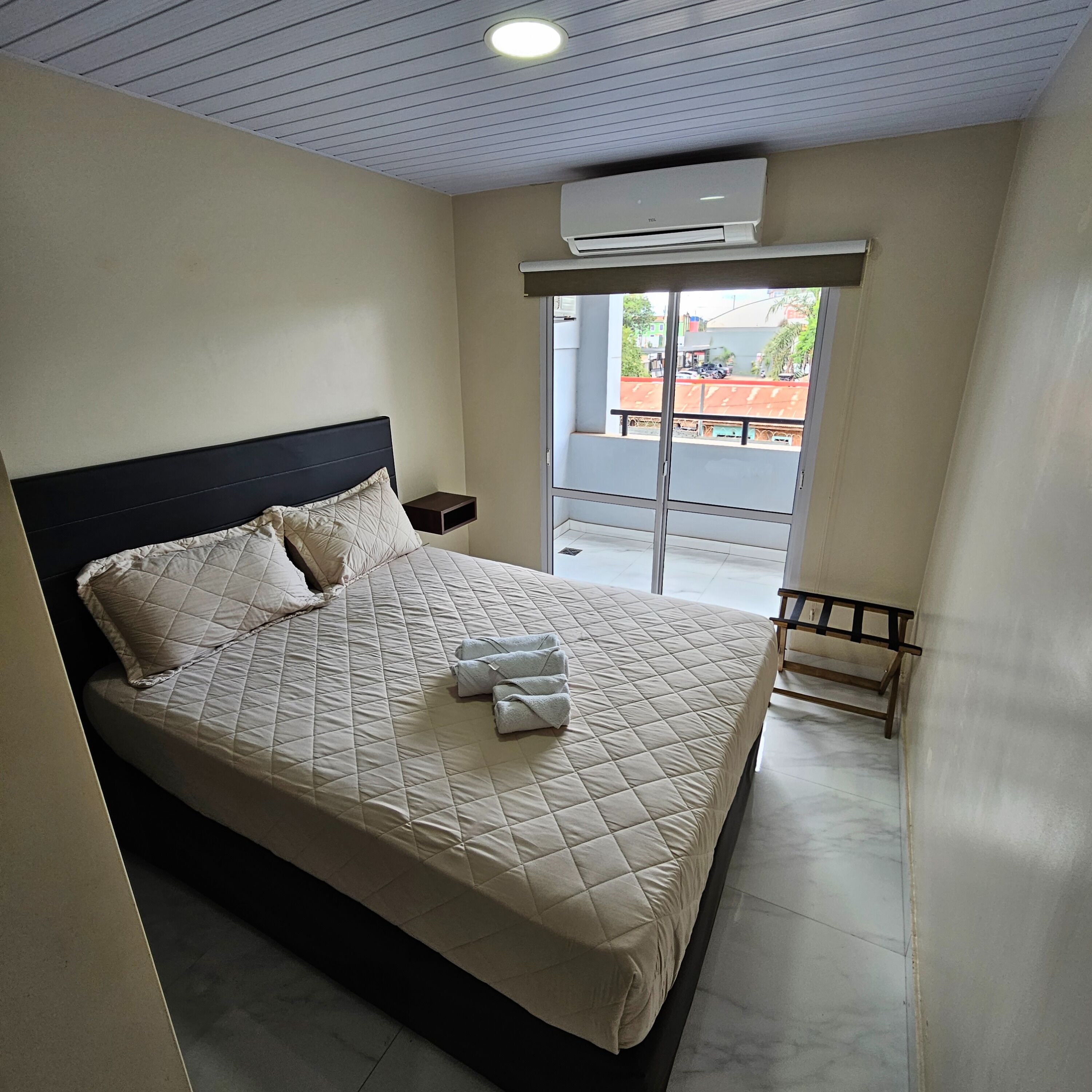 A neatly made bed with a quilted cover, a bedside table, an air conditioning unit, and a view of the outside through a window.