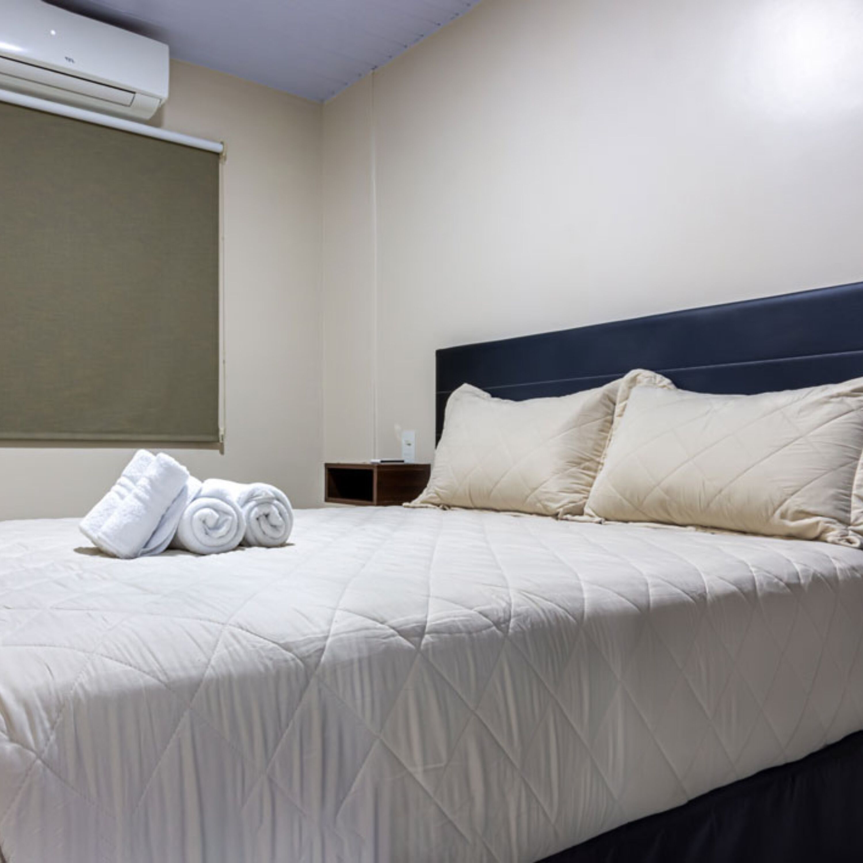 A neatly made bed with rolled towels, a dark headboard, and a plain wall with a window.
