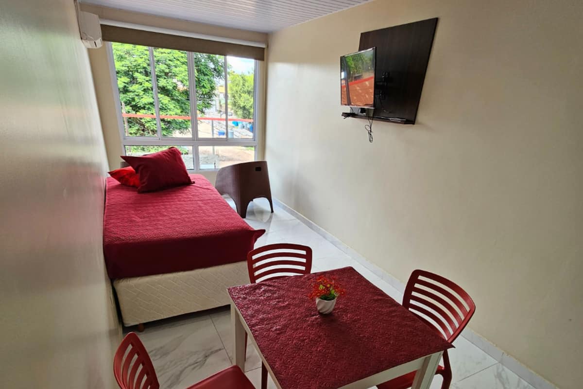Ein Zimmer mit einem Bett, einem Tisch mit Stühlen, einem Fernseher an der Wand und einem Fenster mit Blick auf Bäume.