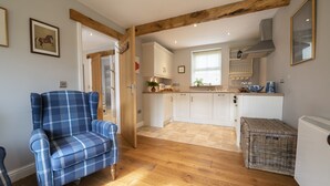 A cozy room with a plaid armchair, wooden flooring, a wicker basket, and a kitchen area with white cabinets and a window. - Saxon House, Harome, North York Moors (Harome)
