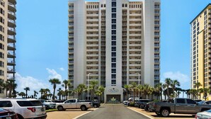 A multi-story hotel with a central entrance, surrounded by parked cars and palm trees. - 🌅Gulf Front|-Beachfront Pool|3BR The Pearl 1201🌅 (Navarre)