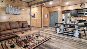 A cozy living room with a brown leather sofa, a wooden coffee table, and a patterned rug. A dining area with a wooden bench and a table set for a meal is visible. The kitchen area includes a refrigerator, microwave, and cabinets. - Modern 4BR Cabin ★ Amazing Location Near Dollywood ★ Grotto Hot Tub ★ Game Room (Sevierville)