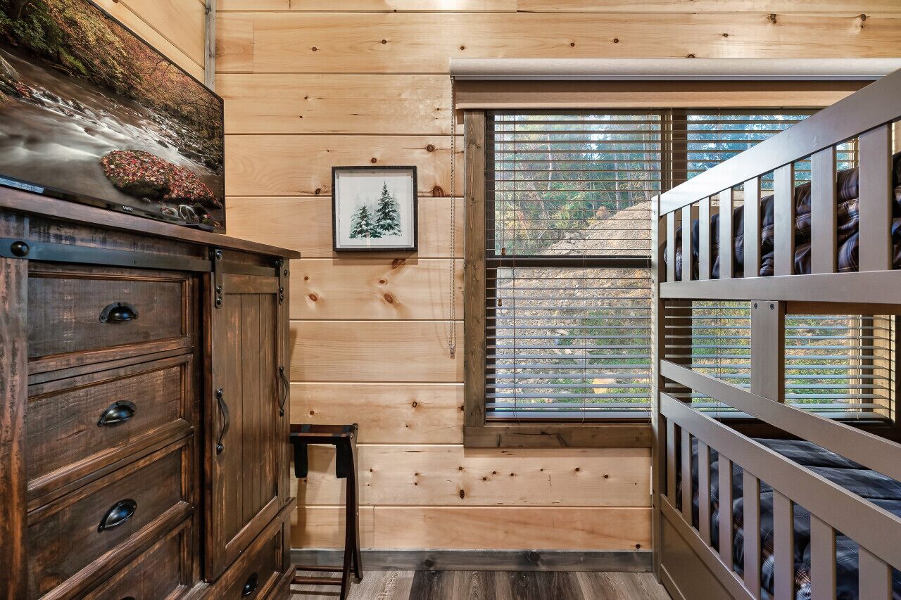Interior de una cabaña de madera con una litera, un aparador, un cuadro enmarcado y una ventana con persianas.