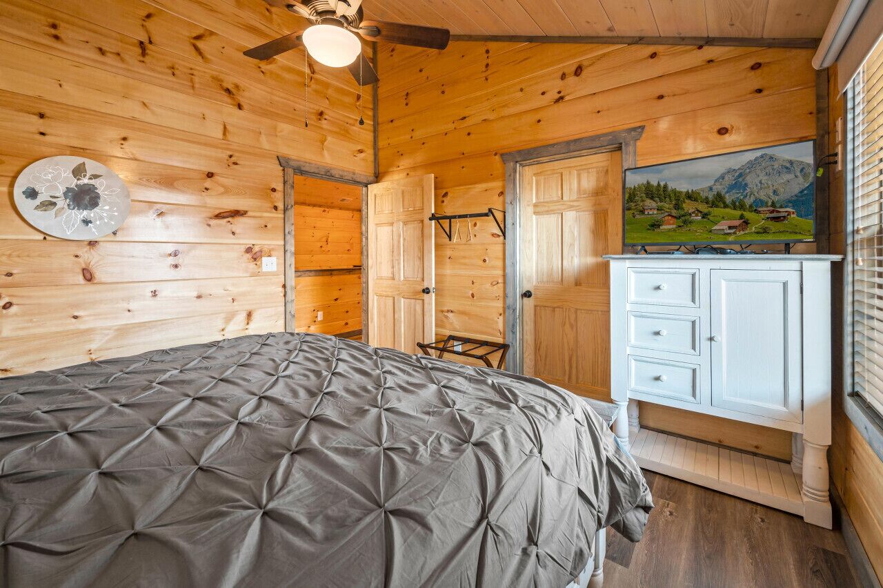 A wooden cabin bedroom with a bed, a dresser, a television, and a painting of a mountain landscape.