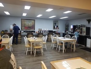 A restaurant with wooden tables and chairs, a counter with a person standing, and a painting on the wall.