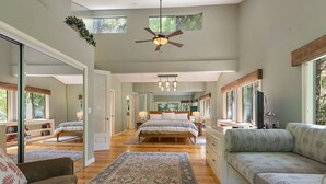 A spacious bedroom with a large bed, a sitting area with a sofa, and a desk. There is a ceiling fan and a window providing natural light.