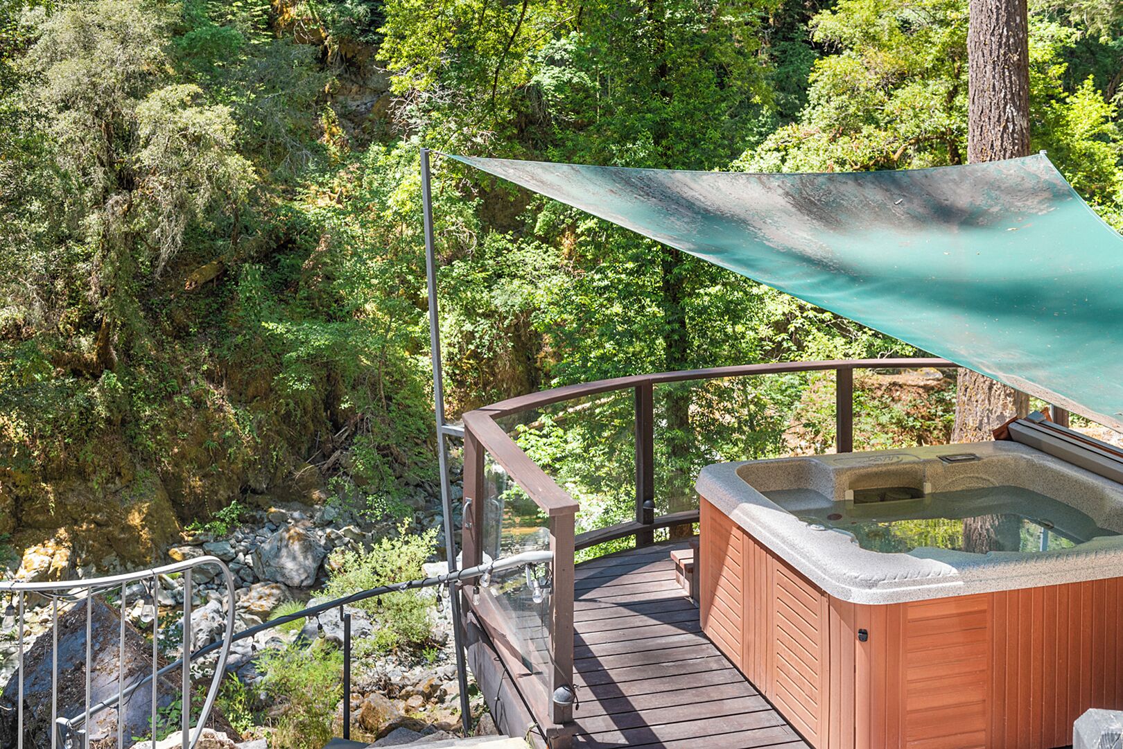 A hot tub on a wooden deck with a metal railing, surrounded by lush greenery and a rocky landscape.