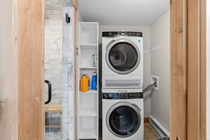 A laundry room with two stacked washing machines, a shower area, and shelves with cleaning supplies.