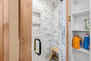 A bathroom with white marble tiles, a wooden bench, and shelves with cleaning supplies.