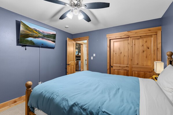 A bedroom with a bed, a ceiling fan, a TV mounted on the wall, and wooden doors.