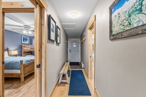 A narrow hallway with a wooden bench, a door at the end, and a bed with a headboard visible on the left.