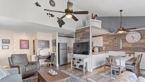 Ein modernes Wohnzimmer mit einer Couch, einem Couchtisch und einem Fernseher. Es gibt einen Essbereich mit Tisch und Stühlen. Der Raum verfügt über einen Deckenventilator und eine Wanduhr.