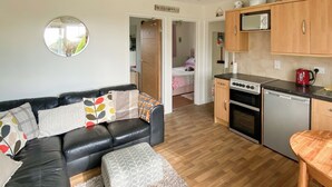 A compact living space with a black leather sofa, a small ottoman, a kitchen area with a stove, microwave, and dishwasher, and a door leading to a bedroom. - Barn Owls Holiday Bungalow (Weston, near Sidmouth)