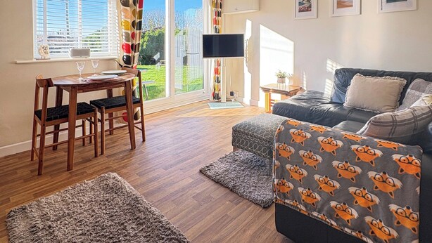 A cozy living room with a wooden floor, a dark grey sofa with a patterned throw, a wooden dining table with chairs, and a large window with a view of the outdoors. - Barn Owls Holiday Bungalow (Weston, near Sidmouth)