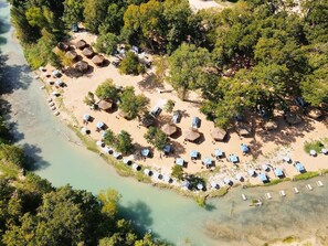 An aerial view of a beach resort with thatched-roof huts and sun loungers arranged in rows along the shoreline. - Son's River Ranch Glamping Cabin #29 (Kingsbury)