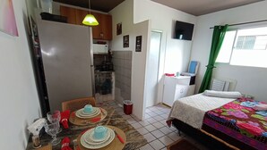 A compact room with a dining table set for two, a bed with a colorful quilt, a TV mounted on the wall, and a small kitchen area. - Lar Recife Olinda Praia 2 (Recife)