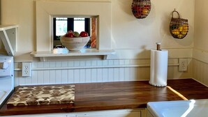 A kitchen with a white countertop, a wooden backsplash, a bowl of fruit on the window sill, and a wall-mounted basket with lemons.