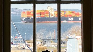 A large cargo ship with multiple containers in the water, viewed through a window with a grid pattern.