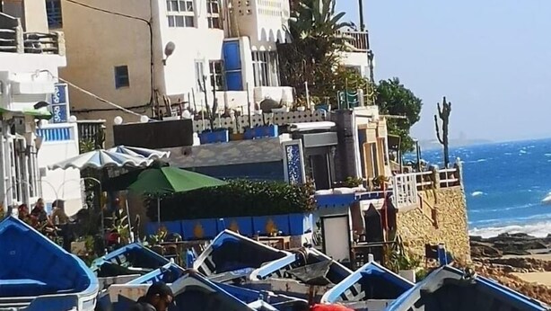 A coastal area with several small boats lined up on the beach, a few people, and buildings in the background.