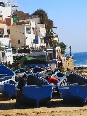 A coastal area with several small boats lined up on the beach, a few people, and buildings in the background.
