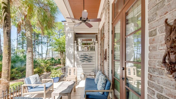 A covered outdoor seating area with wooden furniture, a ceiling fan, and a view of a brick wall and palm trees.