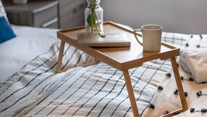 A neatly made bed with a striped bedspread, a wooden bedside table with a book and a mug, a vase with green plants, and a window in the background.