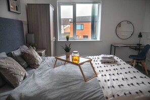 A bedroom with a bed, a desk, a chair, a window with a view of buildings, and a potted plant on the bedside table.
