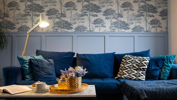 A living room with a blue sofa, a coffee table with a book and a tray, a lamp, and a wall with a tree pattern.