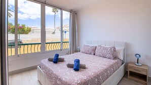 Une chambre avec une grande fenêtre, un lit recouvert d’une literie à motifs, deux serviettes enroulées, une table de chevet avec une lampe, et une vue sur une plage et des palmiers.