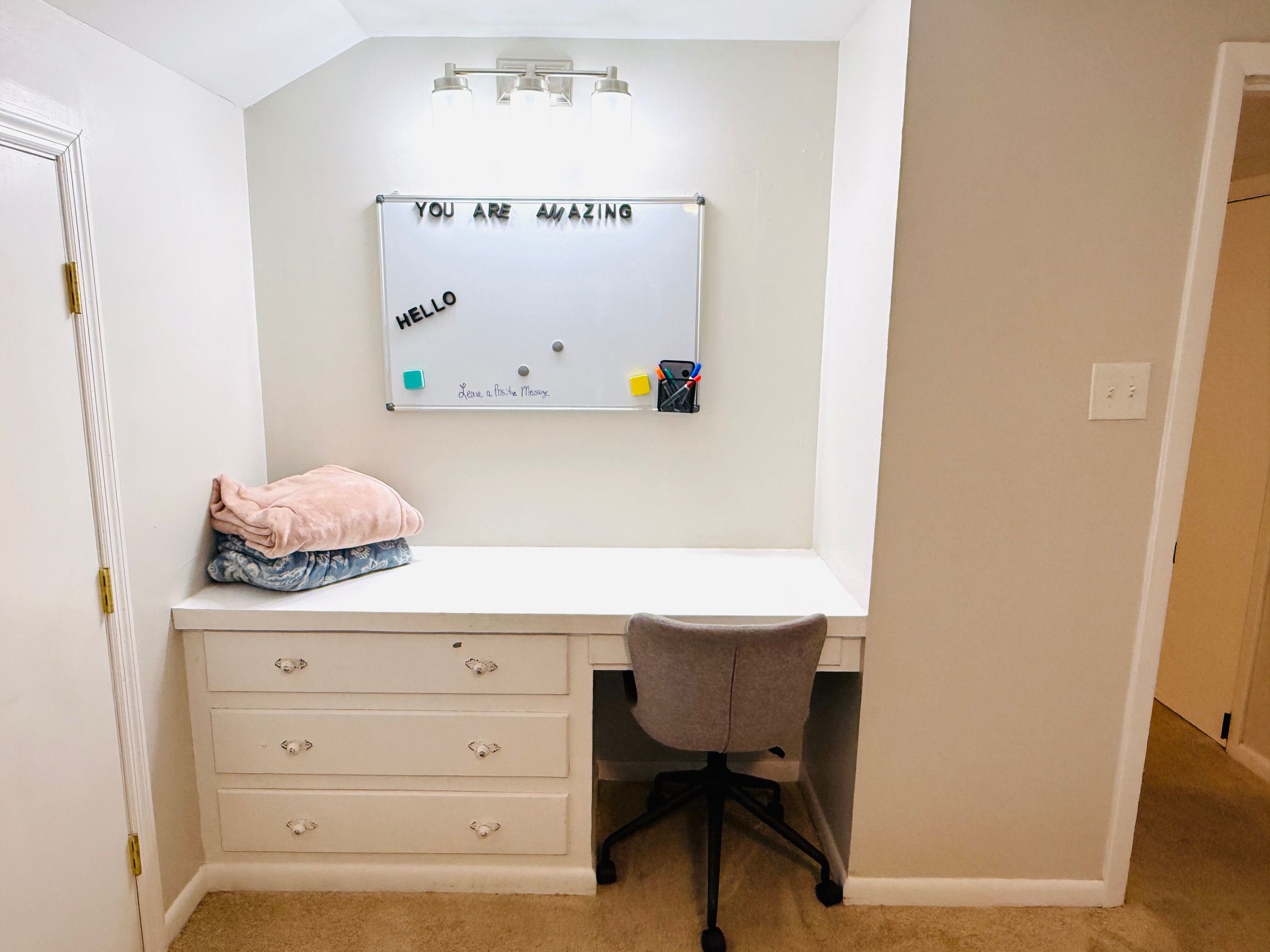 A small, neatly organized room with a white desk, a chair, and a whiteboard above the desk.