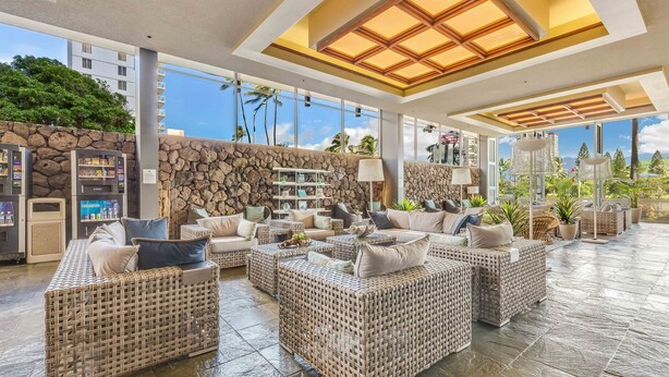 A spacious lounge area with wicker furniture, a stone wall, and large windows offering a view of palm trees and buildings. - 37th FLR Elegant Ocean View Studio|King Bed|Waikiki (Honolulu)