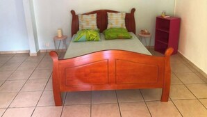 A wooden bed with two pillows, a small bedside table, a pink shelf, and a painting of a pier on the wall. - Studio Cosy, Maisonnette Créole, 100m Jolie Plage (Pointe-à-Pitre)