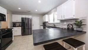 A modern kitchen with white cabinets, a stainless steel refrigerator, a black countertop, and a window with blinds.