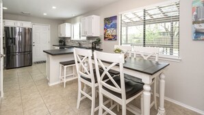 A kitchen with white cabinets, a stainless steel refrigerator, a dark countertop, and a dining area with white chairs.